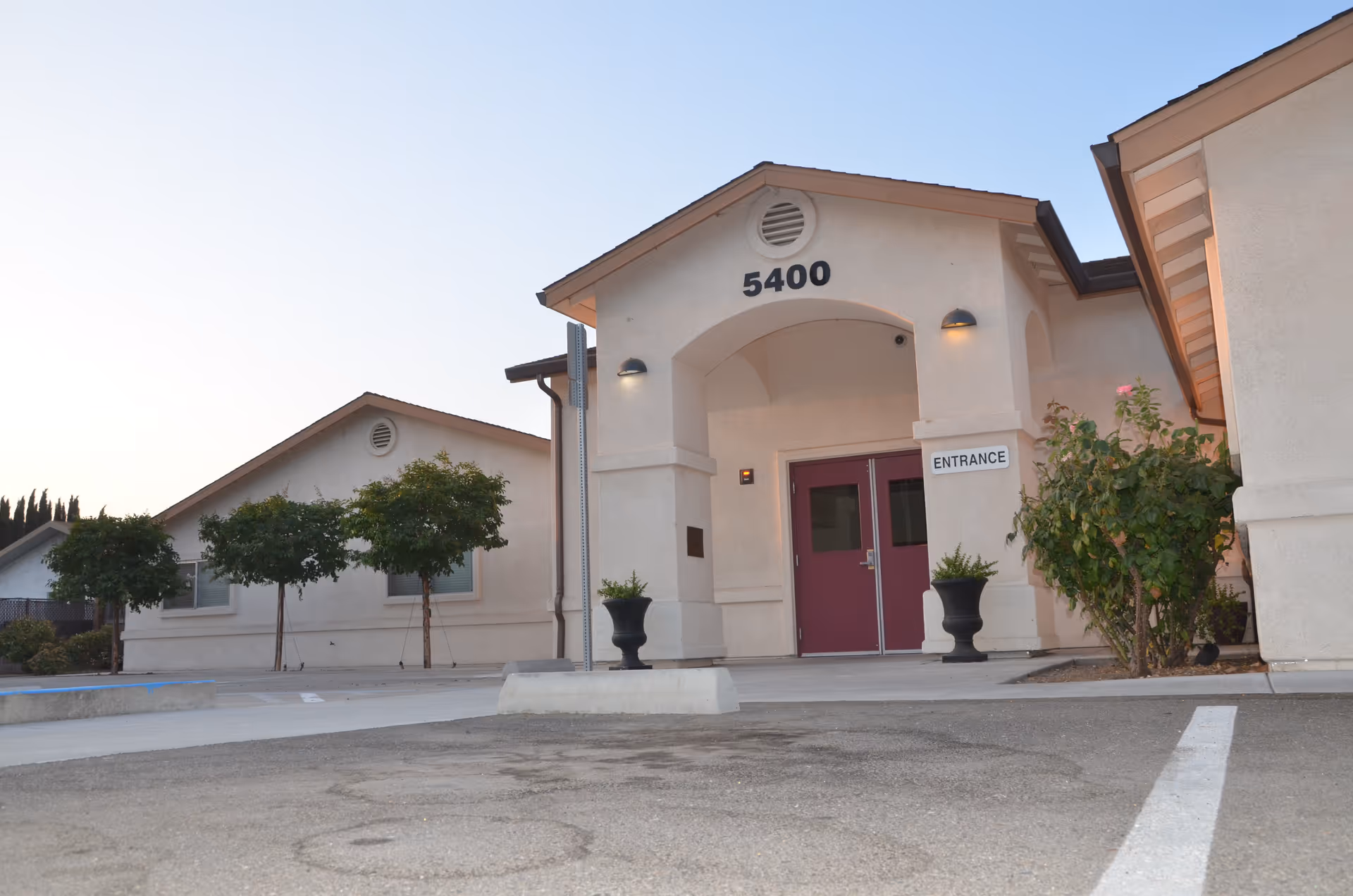 Exterior view of a beige building with the number 5400 above the entrance. The entrance has double doors painted in a reddish color, flanked by two black planters with green plants. There are small trees and bushes around the building, and the photo is taken from a parking lot area with visible parking lines.