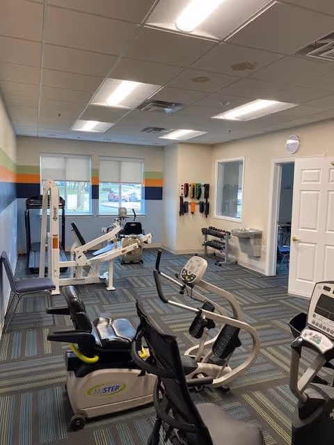 Interior view of a fitness room in a senior living facility with exercise equipment including recumbent bikes, weight machines, and dumbbells. The room has a patterned carpet, white walls with a colorful stripe, fluorescent ceiling lights, and windows with blinds.