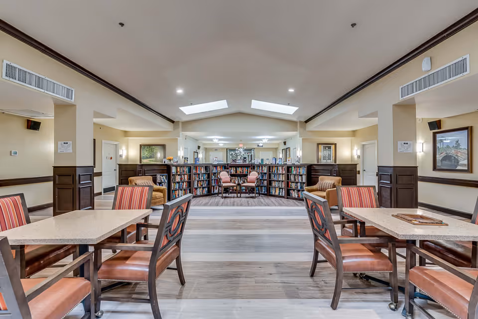 A spacious and well-lit common area in a senior living facility featuring tables with cushioned chairs in the foreground and a semi-circular bookshelf filled with books in the background. Two armchairs and a small table are placed in front of the bookshelf. The room has light-colored walls, wooden paneling, and skylights on the ceiling.