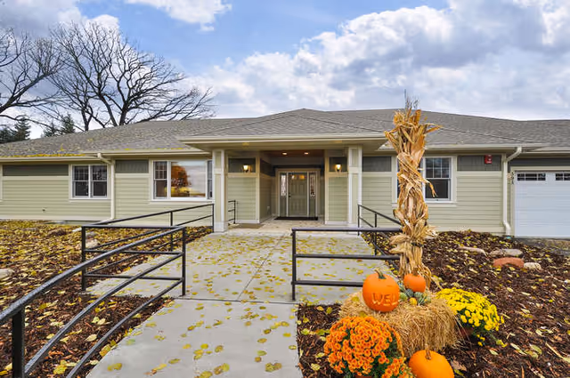 Front exterior view of a single-story building with light green siding and a gray roof. A concrete walkway with black metal railings leads to the entrance, which has double doors with glass panels. The landscaping includes autumn decorations with pumpkins, orange and yellow flowers, and a bundle of dried corn stalks. Trees with bare branches are visible in the background under a partly cloudy sky.