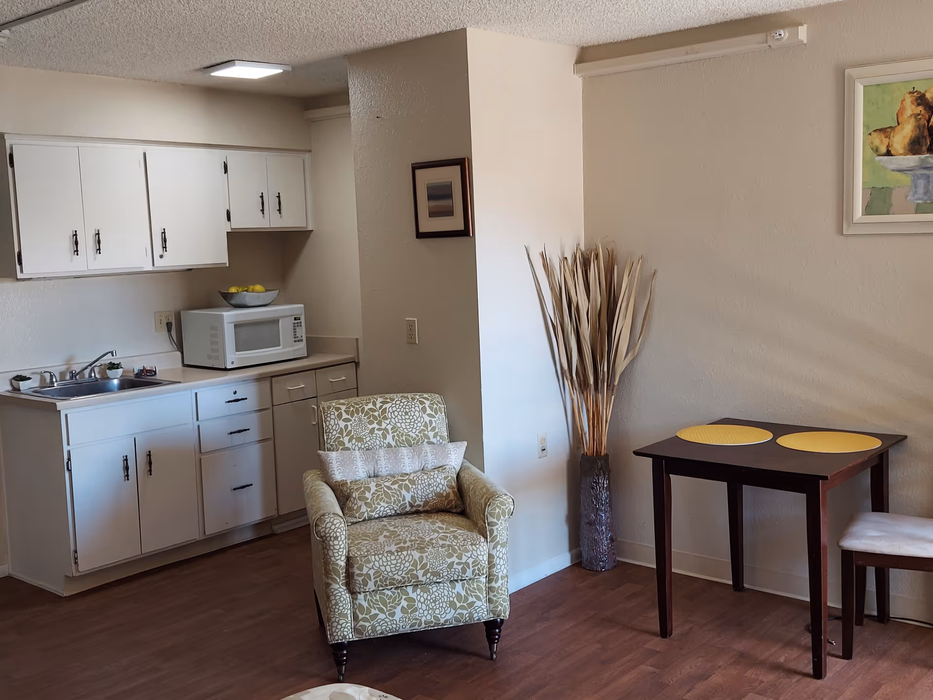 A small living area with a floral patterned armchair, a dark wooden table with two yellow placemats, and a single chair. To the left is a kitchenette with white cabinets, a sink, and a microwave on the counter. The walls are light-colored, and there are two framed pictures and a tall vase with dried decorative plants.