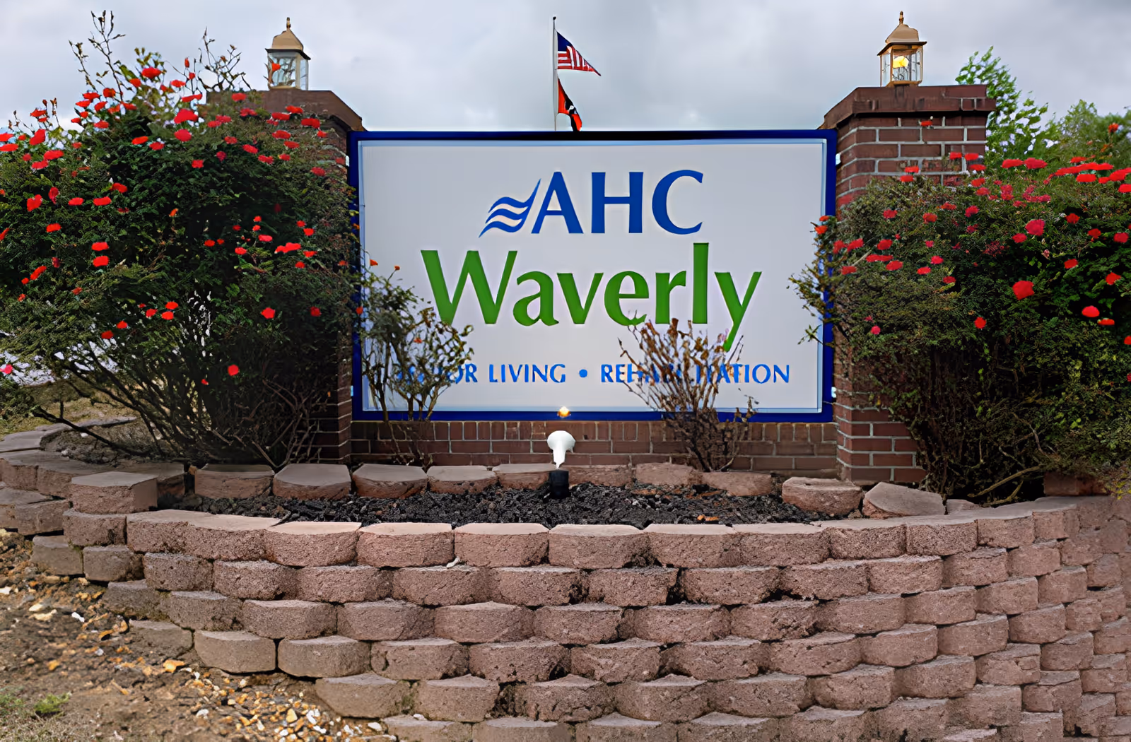 A brick and stone sign for AHC Waverly Senior Living and Rehabilitation, surrounded by red flowering bushes and two lantern-style lights on brick pillars, with an American flag and another flag flying in the background under a cloudy sky.