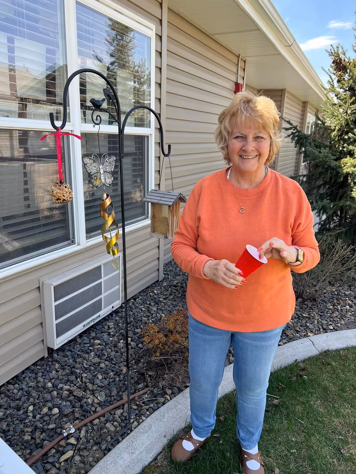 An elderly woman wearing an orange sweatshirt and blue jeans stands outside near a beige building with a window and an air conditioning unit. She is holding a red plastic cup and smiling. There is a metal garden hook with hanging decorations, including a butterfly ornament and a bird feeder, next to her. The ground is covered with rocks and some greenery.