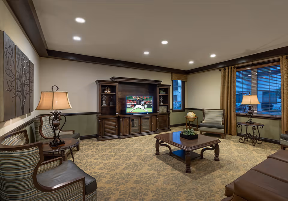 A cozy living room with a patterned carpet, wooden furniture including a coffee table, chairs with striped cushions, and a large entertainment center with a TV showing a baseball game. The room has warm lighting from table lamps, two windows with brown curtains, and decorative wall art featuring tree designs.