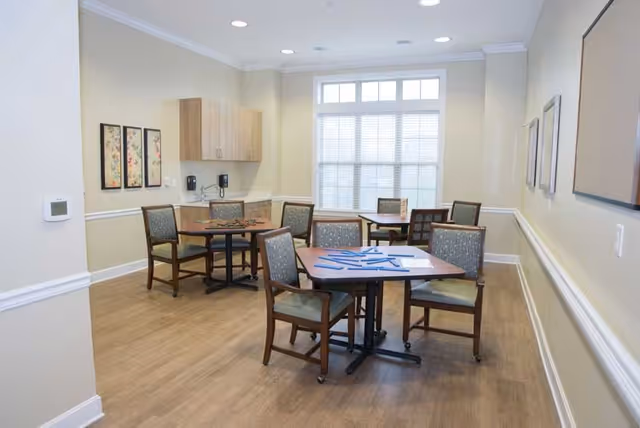 A well-lit room with wooden flooring featuring three square tables, each surrounded by four cushioned chairs. The room has beige walls with white trim, a large window letting in natural light, and framed artwork on the walls. There are cabinets and two wall-mounted telephones on one side of the room.
