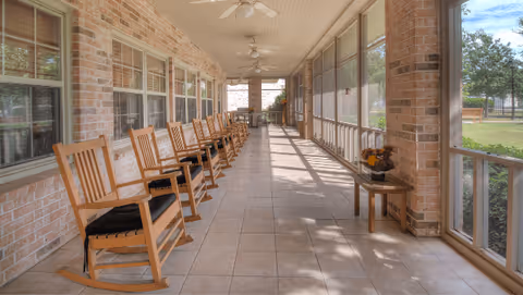 Long screened porch with a row of wooden rocking chairs, tile floor, ceiling fans, and windows overlooking the lawn.