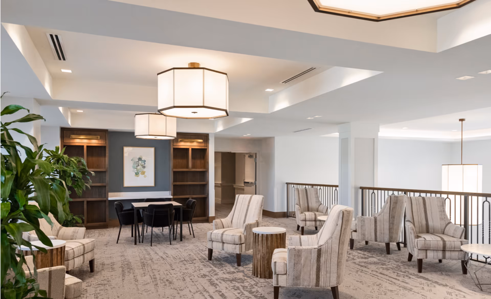 A spacious and well-lit senior living common area with multiple striped armchairs arranged around small round tables. There is a table with four black chairs near a wall with built-in wooden shelves and a framed abstract artwork. Large modern ceiling lights illuminate the room, and a green plant is visible on the left side.