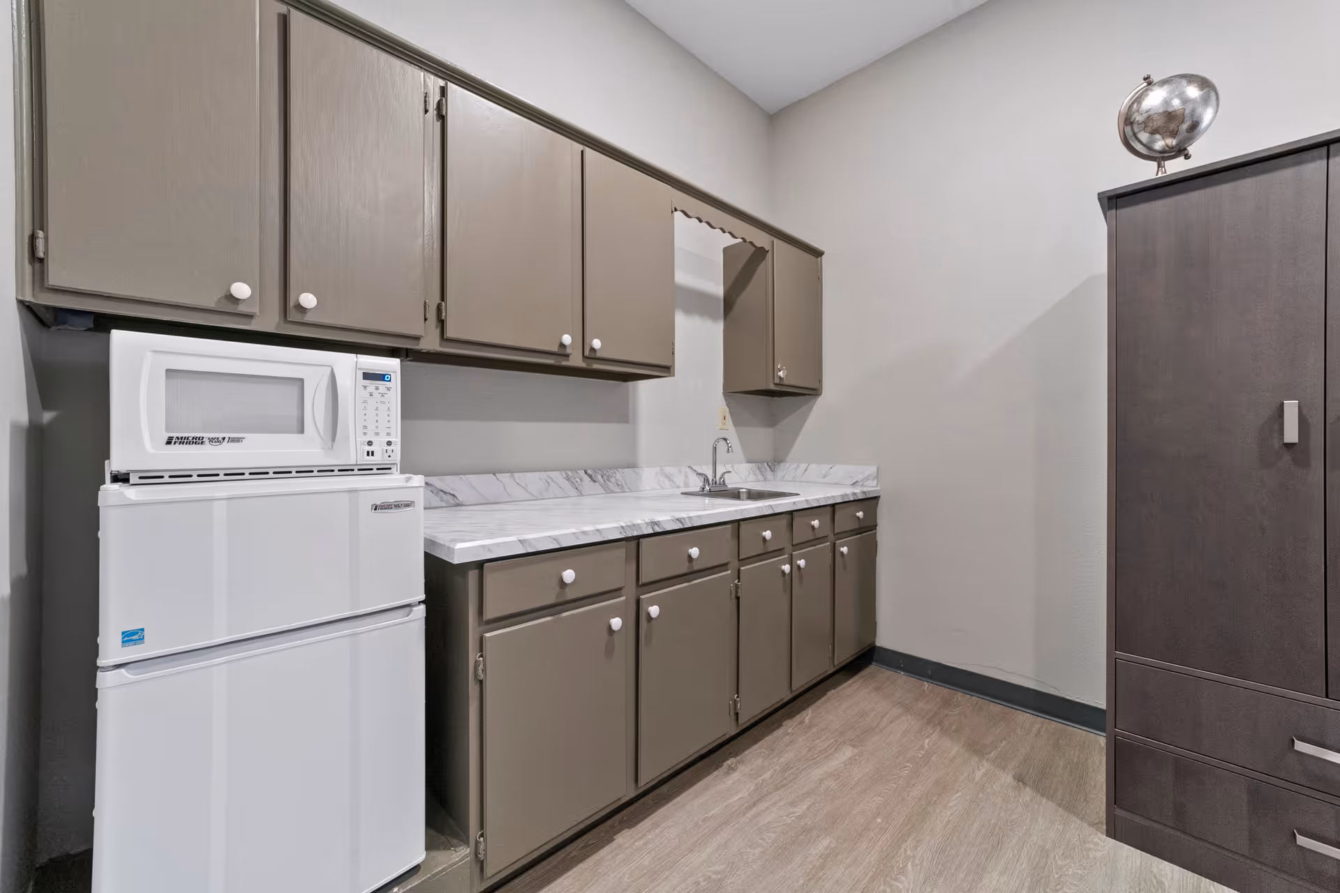 A small kitchen area with gray cabinets, a white microwave on top of a white mini refrigerator, a marble-patterned countertop with a sink, and a dark brown cabinet with a decorative globe on top.