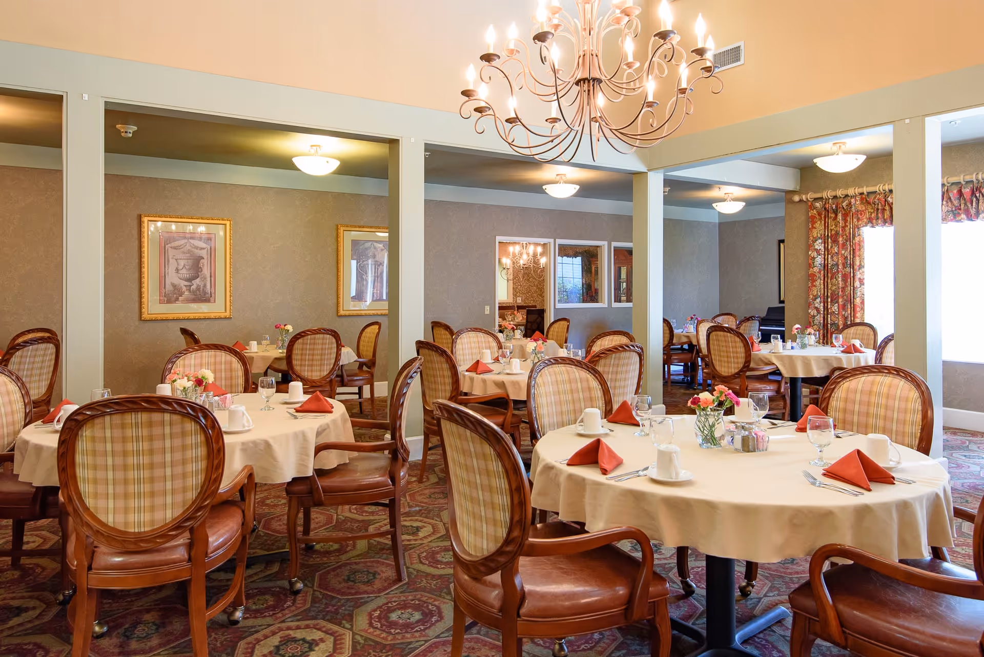 Dining room with round tables set with white tablecloths, red napkins, floral centerpieces, and upholstered chairs under chandeliers.