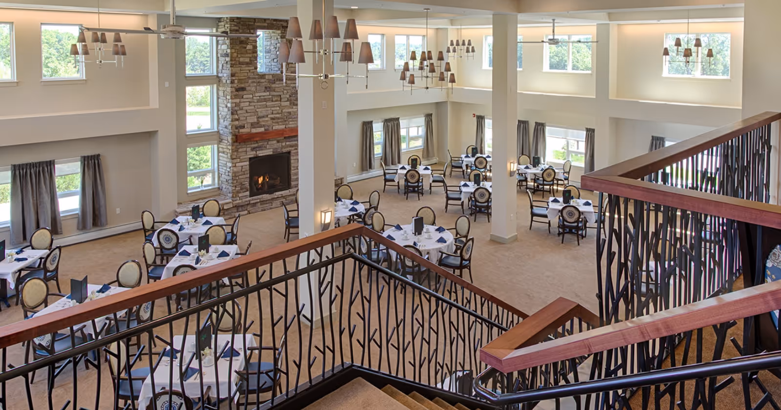 Two-story dining room with round tables set for service, decorative staircase railing, chandeliers, and a stone fireplace.