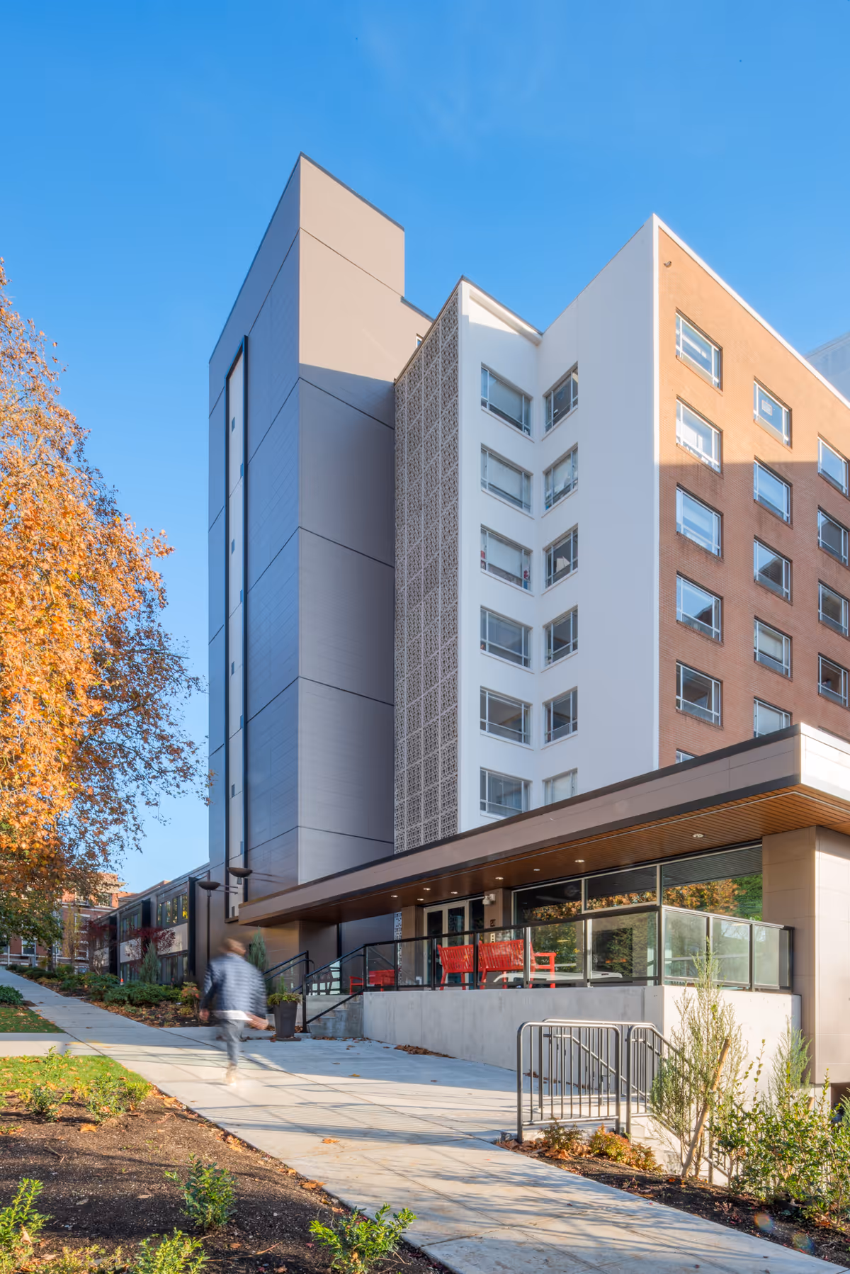 Exterior view of a modern multi-story building with a sidewalk leading up to the entrance. The building has a mix of white, gray, and brick facades with large windows. There is a person walking on the sidewalk and some autumn-colored trees on the left side.