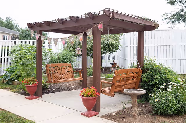 A wooden pergola with two wooden swings hanging from it, situated on a concrete patio surrounded by garden beds with green plants and flowers. There are two red flower pots with blooming flowers placed at the base of the pergola posts. A white fence encloses the garden area.
