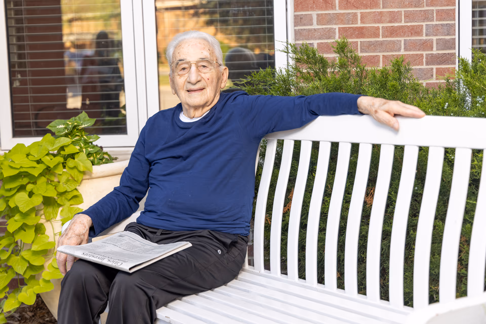An elderly man wearing glasses and a navy blue long-sleeve shirt sits on a white bench outdoors, holding a newspaper. Behind him are green plants, a brick wall, and a window with blinds.