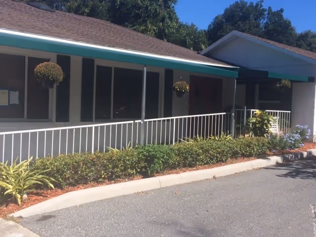 Front exterior of a single-story building with green awnings, a covered walkway, white ramp railing, hanging planters, and landscaped curbside.