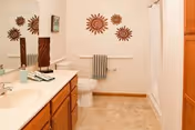 A clean bathroom with a single sink vanity featuring wooden cabinets, a large mirror, a toilet, and a bathtub with a white shower curtain. The walls are decorated with three sun-shaped wall hangings above the toilet, and a towel is hanging on a rack below them.