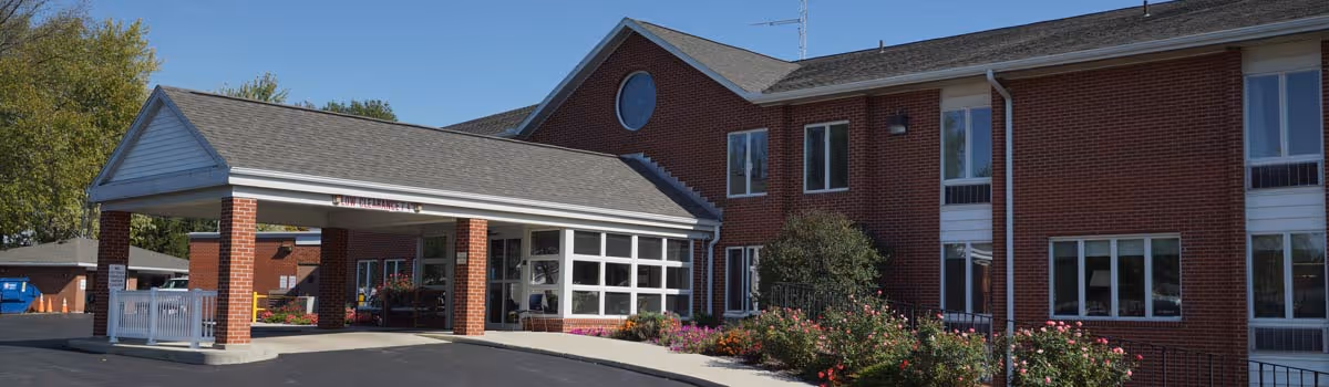 Exterior view of a two-story brick building with a covered entrance and a driveway. There are windows on both floors and a garden with bushes and flowers in front of the building under a clear blue sky.