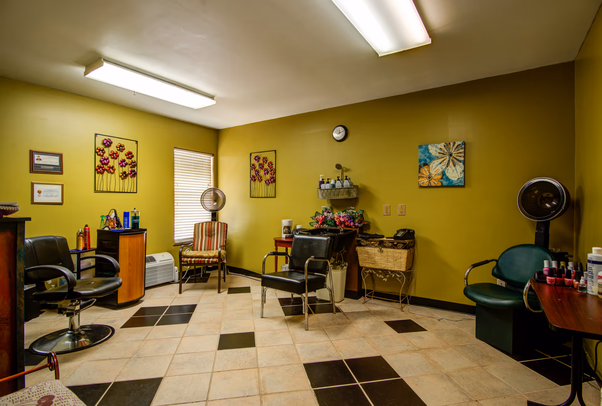 Interior of a salon room with green walls and tiled floor. The room contains salon chairs, a hair dryer, a small table with nail polish bottles, and decorative wall art including flower designs and a clock. There is a window with blinds and various salon supplies on shelves and tables.