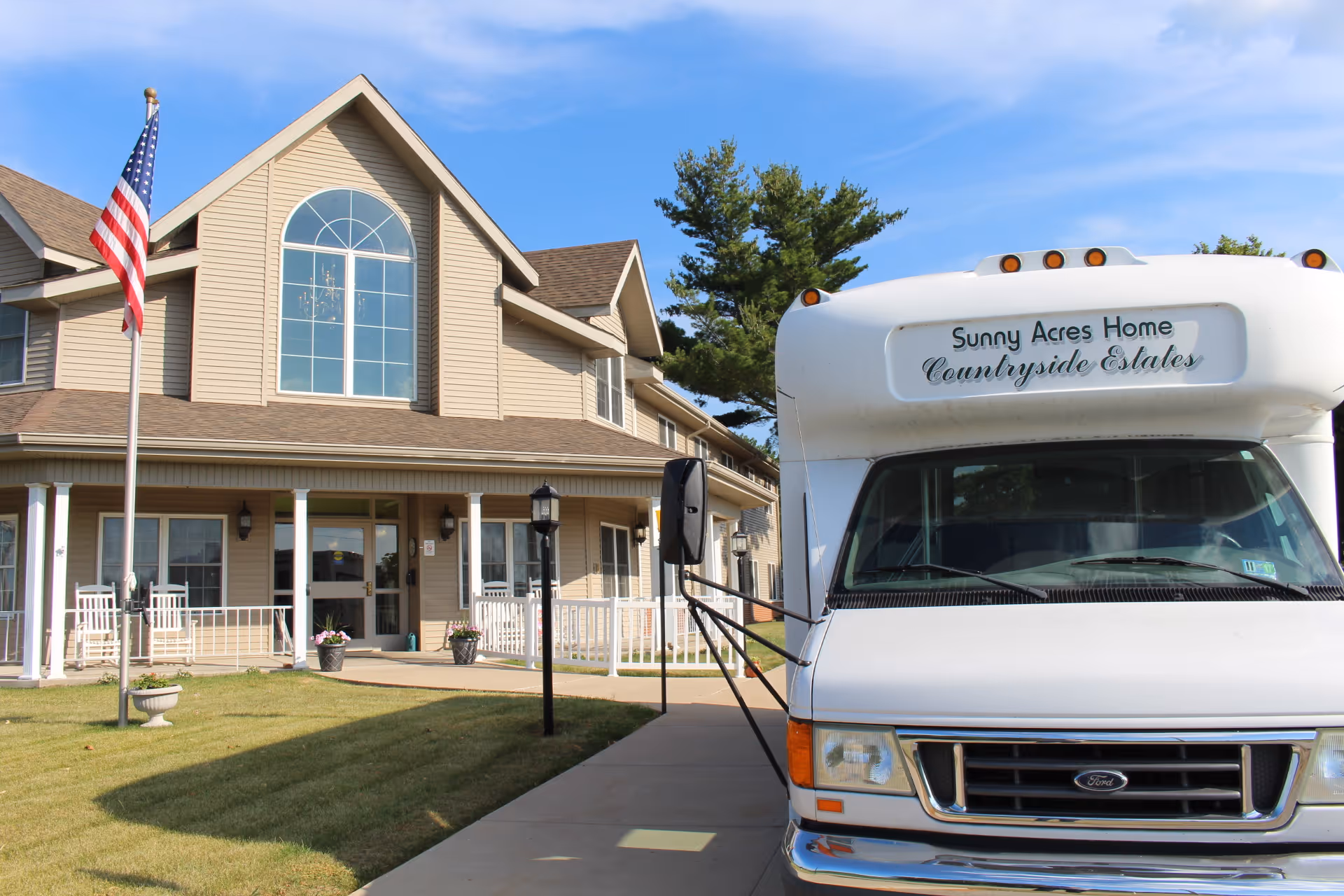 Exterior view of Sunny Acres Nursing Home building with beige siding, large arched window, and a porch with white rocking chairs. An American flag is on a flagpole in the front yard. A white shuttle bus with the sign 'Sunny Acres Home Countryside Estates' is parked on the driveway.
