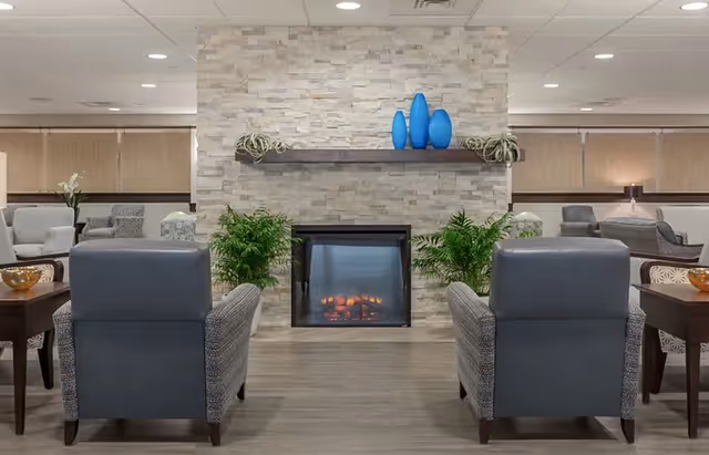 A cozy lounge area with two gray armchairs facing a modern stone fireplace. The fireplace has a wooden mantel decorated with three blue vases and two small plants. There are additional seating arrangements with chairs and tables in the background, and the room has a neutral color palette with soft lighting.