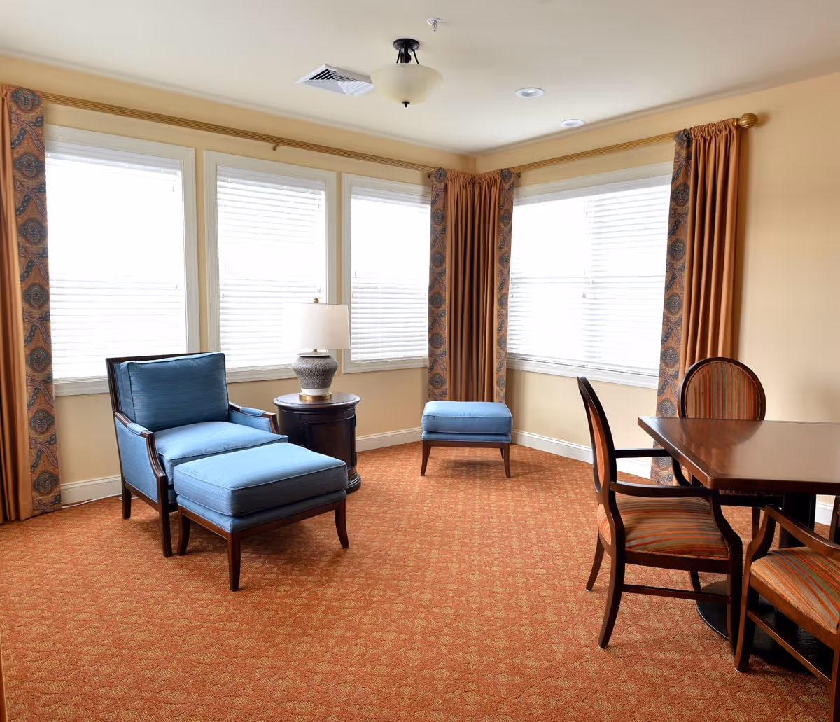 A cozy sitting area with a blue cushioned armchair and matching ottoman, a small round wooden side table with a lamp, and a wooden dining table with three striped cushioned chairs. The room has large windows with white blinds and patterned brown curtains, beige walls, and an orange patterned carpet.