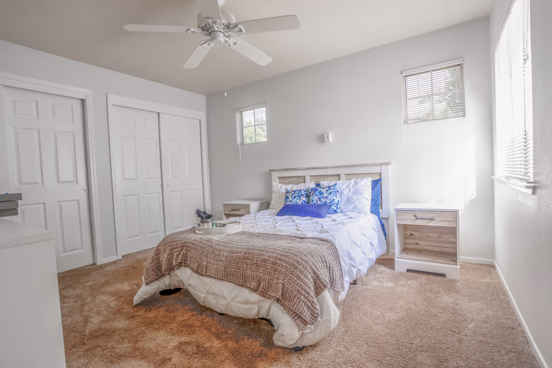 A bright bedroom with a bed centered against a white wall, featuring white and blue bedding and a brown throw blanket. There are two small windows with blinds on the wall behind the bed, a ceiling fan above, beige carpet flooring, two nightstands on either side of the bed, and a set of white double closet doors on the left side of the room.