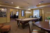 A cozy indoor recreational room featuring a pool table with cues and balls set up, a round table with chairs in the foreground, comfortable seating including armchairs near windows with curtains, and warm lighting from ceiling fixtures.