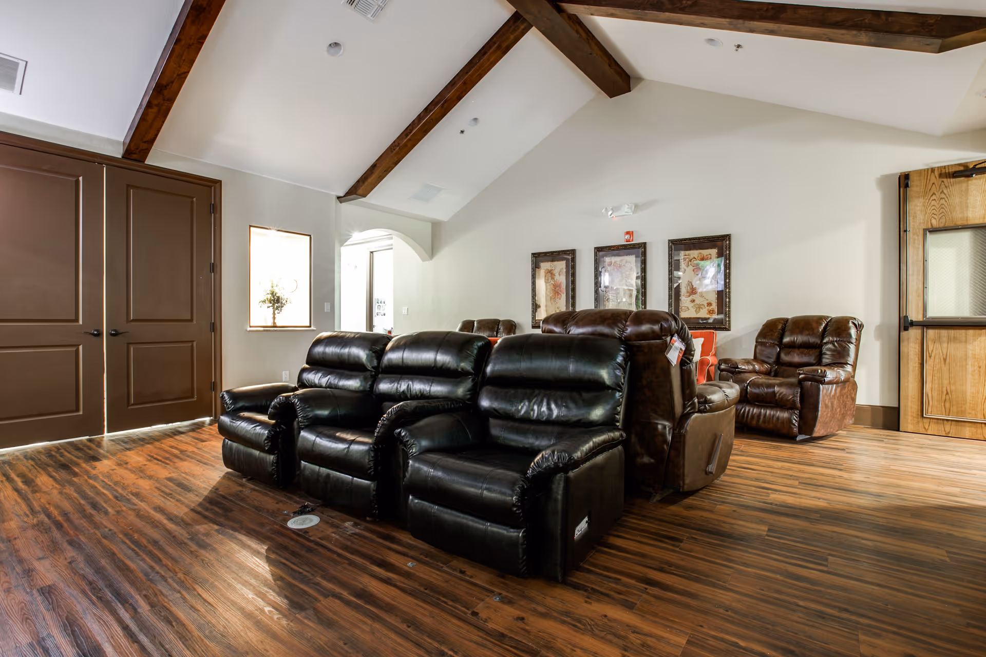 Interior view of a living room area in Avalon Memory Care featuring several dark leather recliners arranged on a wooden floor under a vaulted ceiling with exposed wooden beams. The walls are light-colored with framed artwork and a wooden door is visible on the right side.