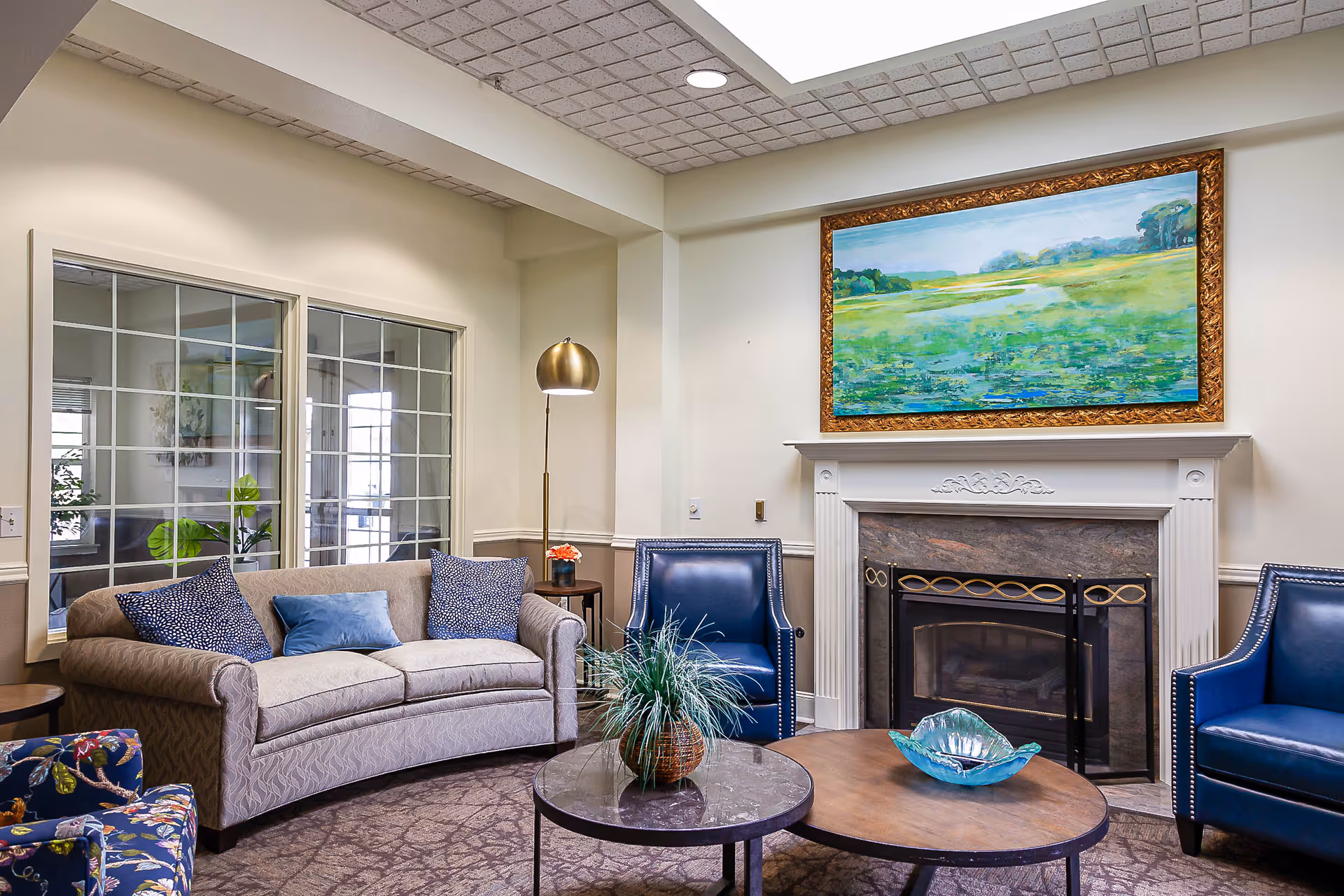 A cozy living room area with a beige curved sofa adorned with blue and patterned cushions, two blue leather armchairs, a floral patterned chair, and two round coffee tables. A decorative plant and a blue glass bowl are on the tables. The room features a white fireplace with a black and gold screen, above which hangs a large framed painting of a green landscape. A tall brass floor lamp stands next to the sofa, and a large window with grid panes is visible on the left wall.