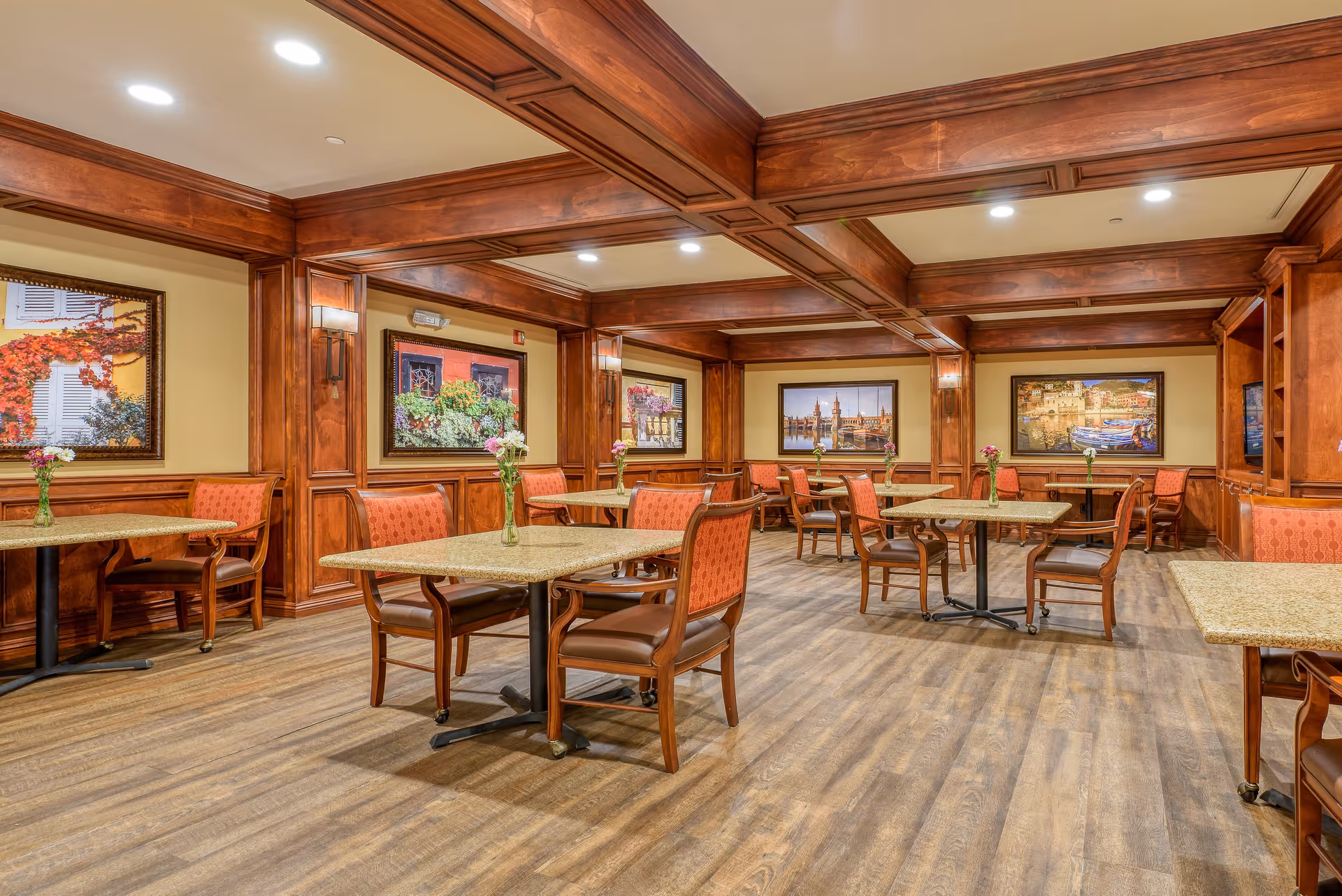 A dining room with wooden paneling and beams on the ceiling, several tables with granite tops and wooden chairs with orange upholstery. Each table has a small vase with flowers. The walls are decorated with framed pictures of scenic outdoor views.