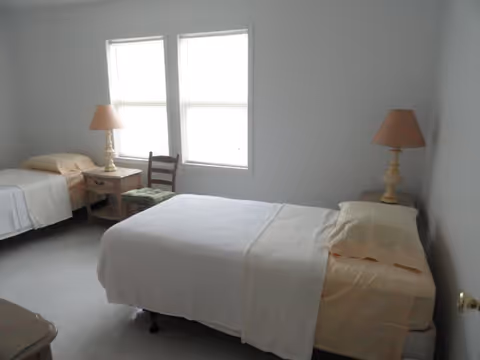 A simple bedroom with two single beds, each with light-colored bedding and a pillow. There are two bedside tables, each with a lamp. Between the beds is a wooden chair with a cushion. Two windows with closed blinds are on the wall behind the chair.