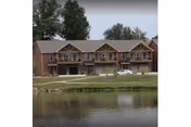 View of a two-story brick residential building with balconies overlooking a pond, surrounded by trees and a grassy area.