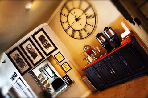 Interior view of a reception or common area with a large wall clock, a dark wooden cabinet holding beverage dispensers and cups, framed black and white photos on the wall, and a reception desk with a chair visible in the background.