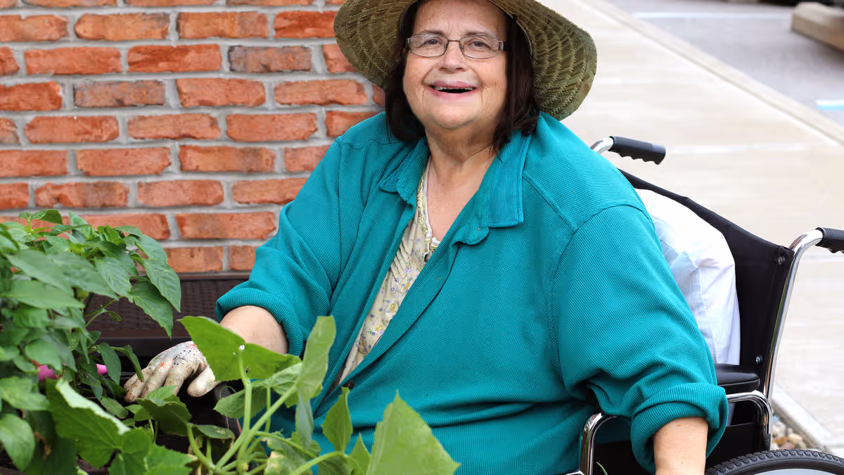 An elderly woman wearing a green jacket and a wide-brimmed hat is sitting in a wheelchair outdoors next to a brick wall. She is smiling and appears to be gardening, with green plants and leaves visible in front of her.