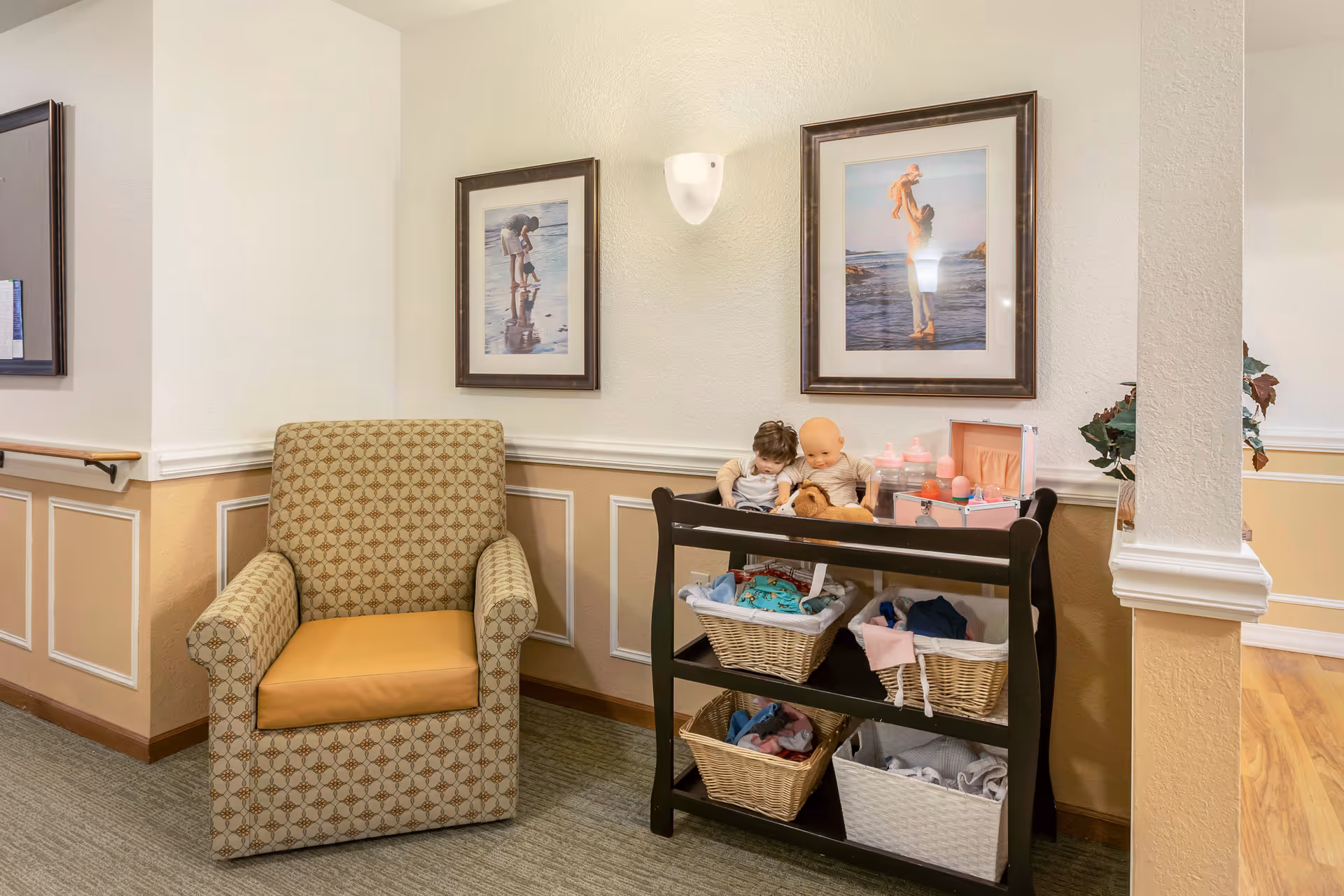 A cozy corner in a senior living facility with a patterned armchair next to a black shelving unit holding dolls, baby bottles, and baskets filled with clothes. Two framed pictures of adults playing with children at the beach hang on the wall above the shelving unit. The walls are painted beige and white with decorative molding, and there is a wall-mounted light fixture between the pictures.