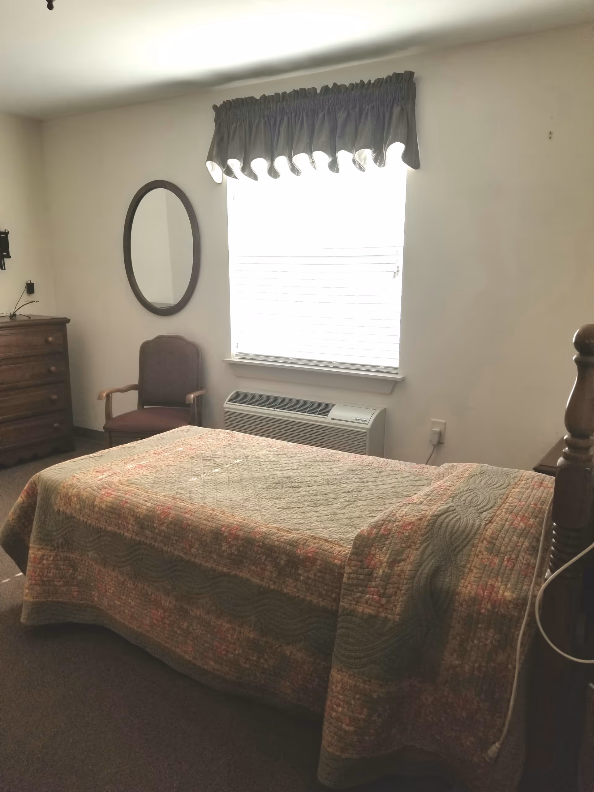 A small bedroom with a single bed covered with a quilted bedspread. There is a wooden dresser with drawers on the left side, a wooden chair with armrests next to it, and an oval mirror hanging on the wall above the chair. A window with blinds and a dark valance is centered on the wall, with an air conditioning unit installed below it. The room has plain white walls and a carpeted floor.