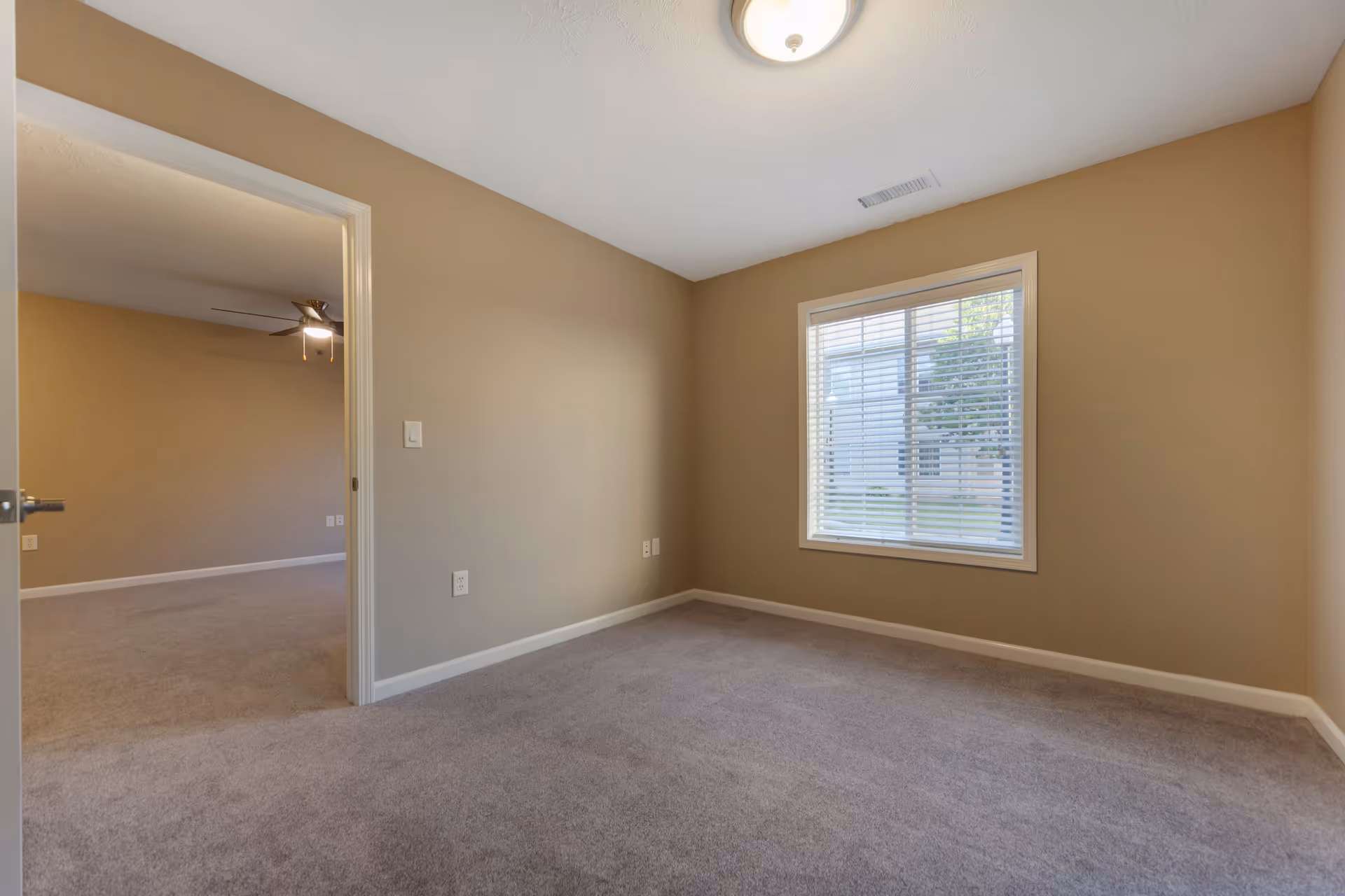 Empty room with beige walls and gray carpet, a window with white blinds letting in natural light, and an open doorway leading to another room with a ceiling fan and light.
