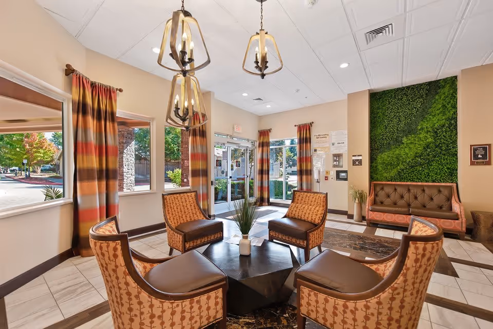 A bright and welcoming senior living facility lobby with four patterned armchairs arranged around a square black coffee table with a small plant. The room features large windows with colorful striped curtains, three hanging pendant lights, a green vertical garden wall, and a brown tufted bench. The entrance door is visible with natural light coming through.