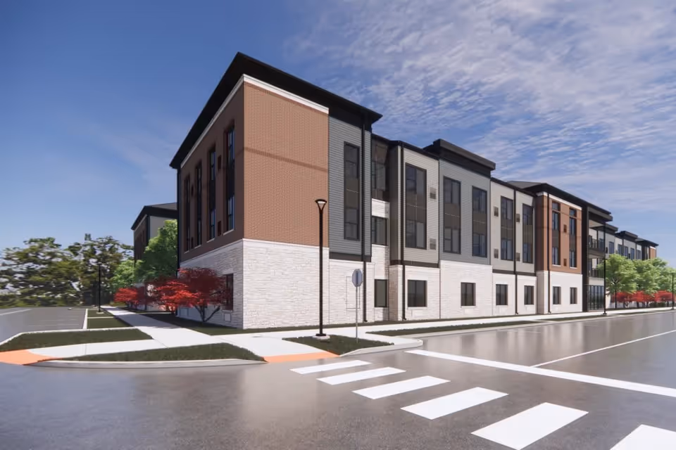 Three-story modern senior living apartment building on a street corner with a crosswalk, sidewalk and landscaped trees under a partly cloudy sky.