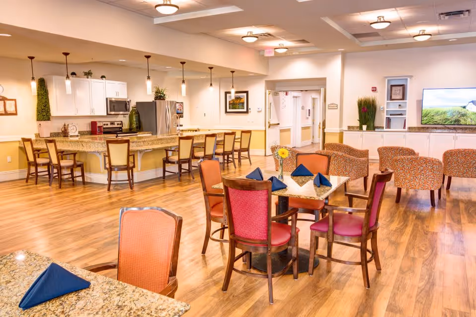 A bright and spacious common area in a senior living facility featuring a kitchen with a granite countertop island and bar stools, several dining tables with chairs, and a seating area with patterned armchairs. The room has wooden flooring, pendant lights over the kitchen island, and a flat-screen TV mounted on the wall.