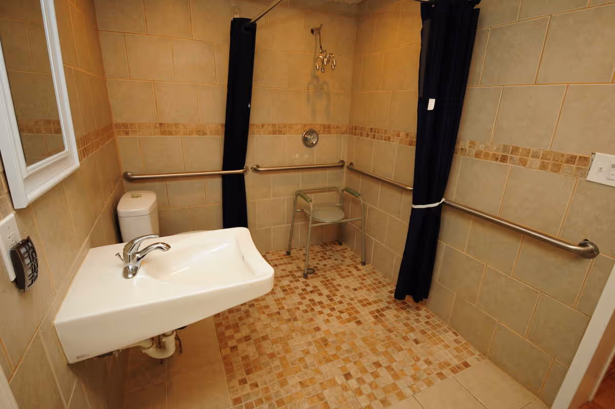Accessible bathroom with beige tiled walls and floor, featuring a wall-mounted sink, a toilet, grab bars along the walls, a shower area with a shower chair, and a dark shower curtain.
