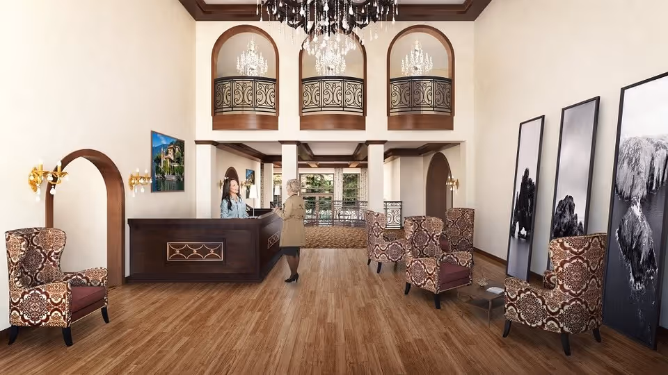 A spacious and elegant reception area with wooden flooring, patterned armchairs, a dark wooden reception desk with a receptionist and a visitor, large black and white framed photographs leaning against the wall, ornate chandeliers hanging from the ceiling, and decorative wrought iron balcony railings on the upper level.