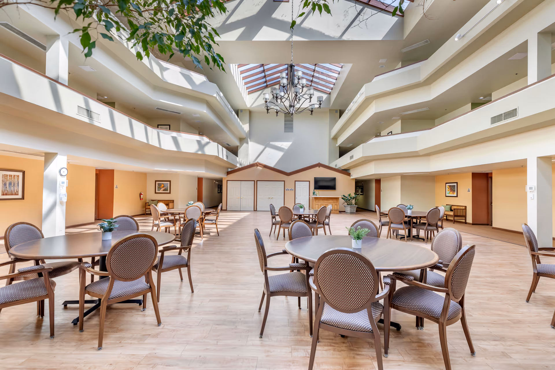 A spacious, well-lit common area in a senior living facility with multiple round tables and chairs arranged on a wooden floor. The room features a high ceiling with a large skylight and a chandelier, allowing natural light to fill the space. There are balconies on the upper floors overlooking the area, and the walls are decorated with framed artwork. A TV is mounted on the far wall above a wooden cabinet, and potted plants add greenery to the room.