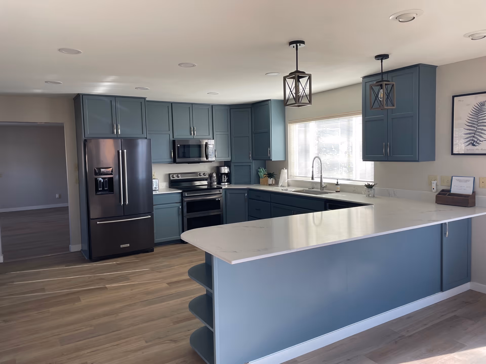 Modern kitchen with blue cabinets, stainless steel refrigerator, oven, microwave, and a large white countertop island. Two pendant lights hang from the ceiling above the island, and a window with blinds is above the sink. The floor is light wood, and there is a doorway leading to another room.