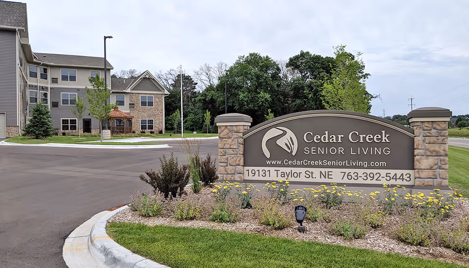 Entrance sign for Cedar Creek Senior Living with the building and landscaped grounds in the background under a cloudy sky.