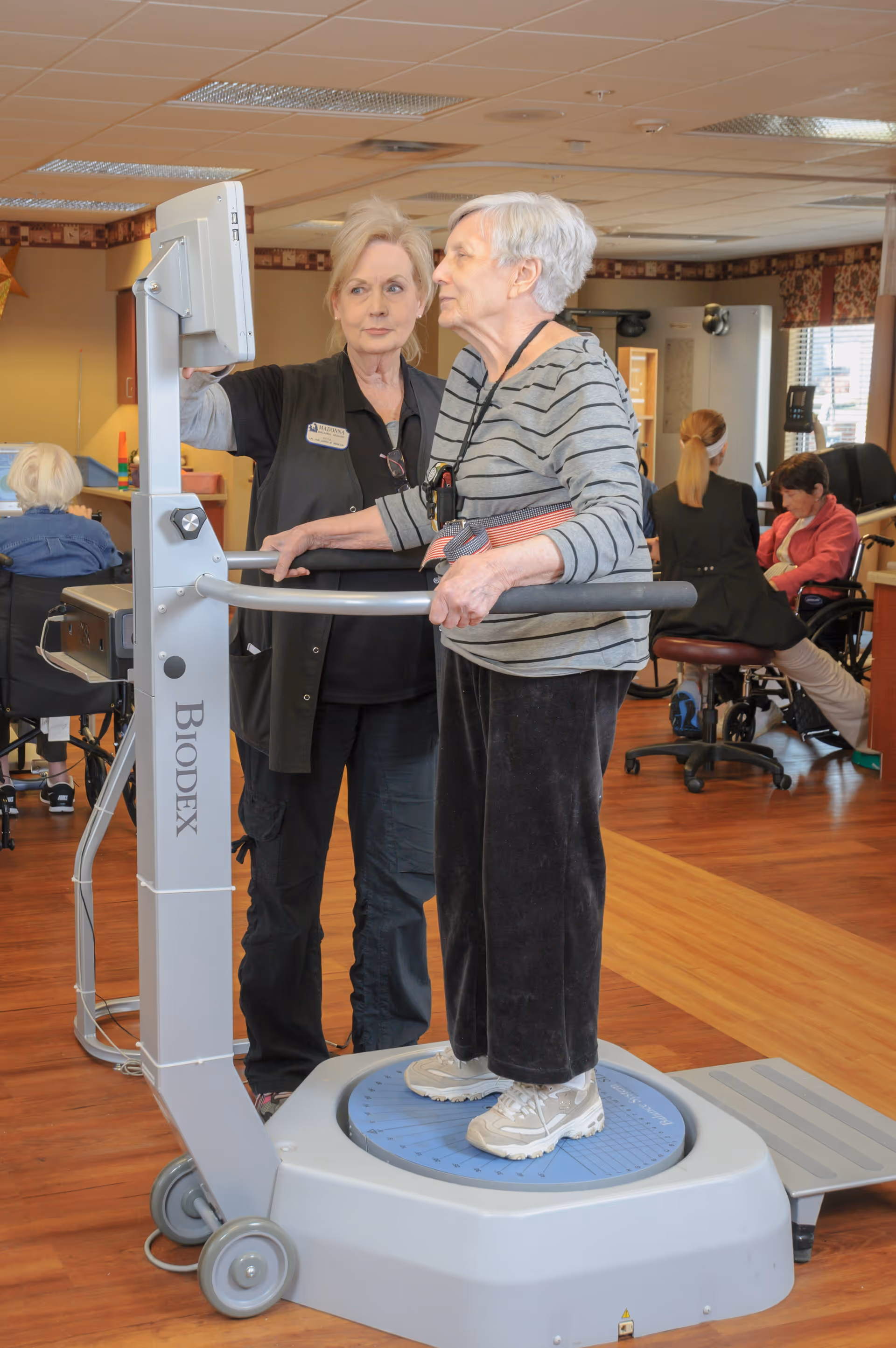 An elderly woman stands on a Biodex balance testing machine while a staff member assists her by operating the machine's monitor in a senior living facility common area. Other elderly residents and staff are visible in the background, some seated and engaged in activities.