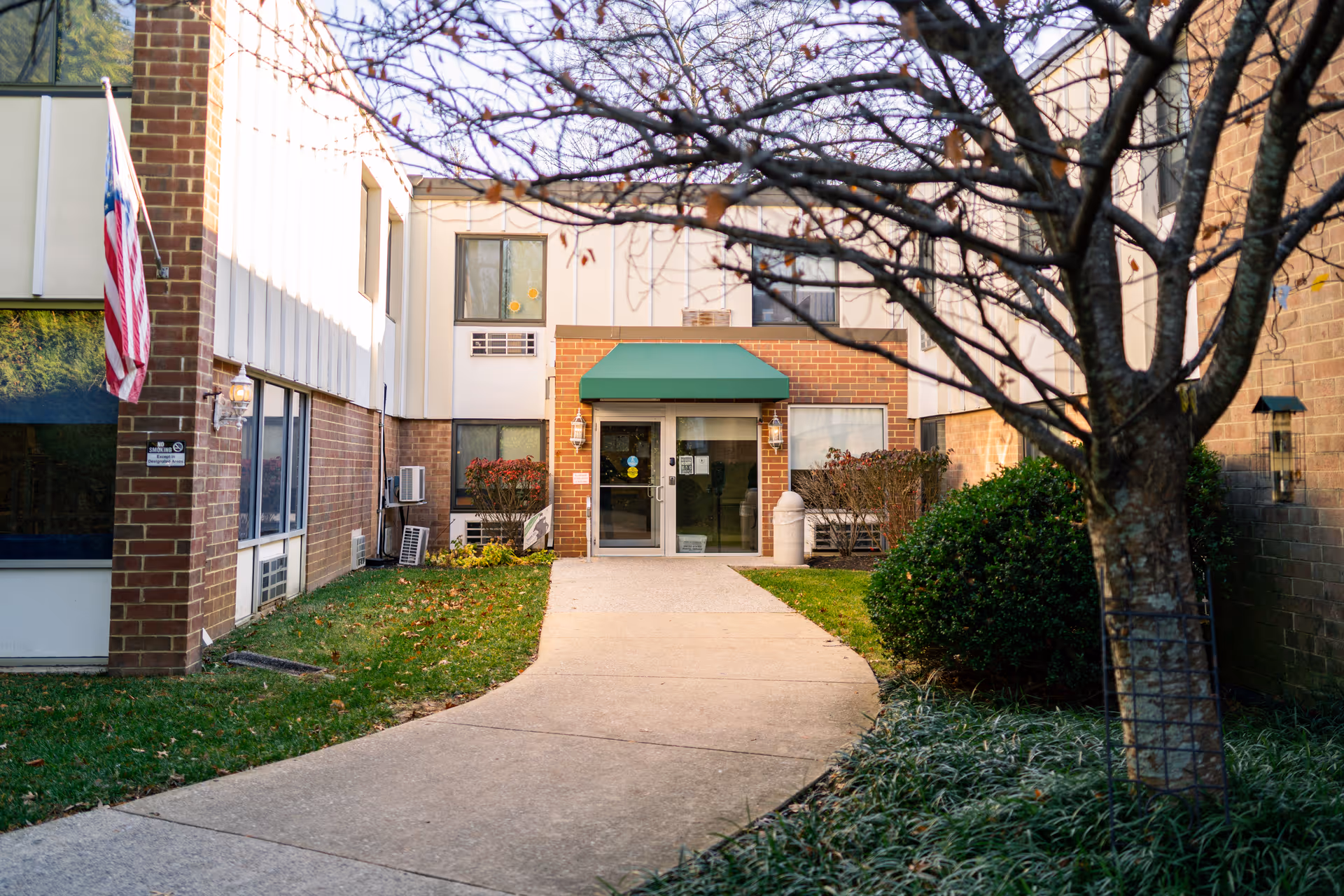 Exterior view of Autumn Lake Healthcare at Catonsville showing the entrance with a green awning above the door, a concrete pathway leading to the entrance, an American flag on the left side, and trees and bushes surrounding the walkway.