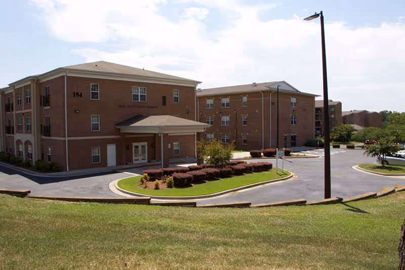 Front exterior of a three-story brick senior living building with a covered entrance, circular driveway, and landscaped lawn.