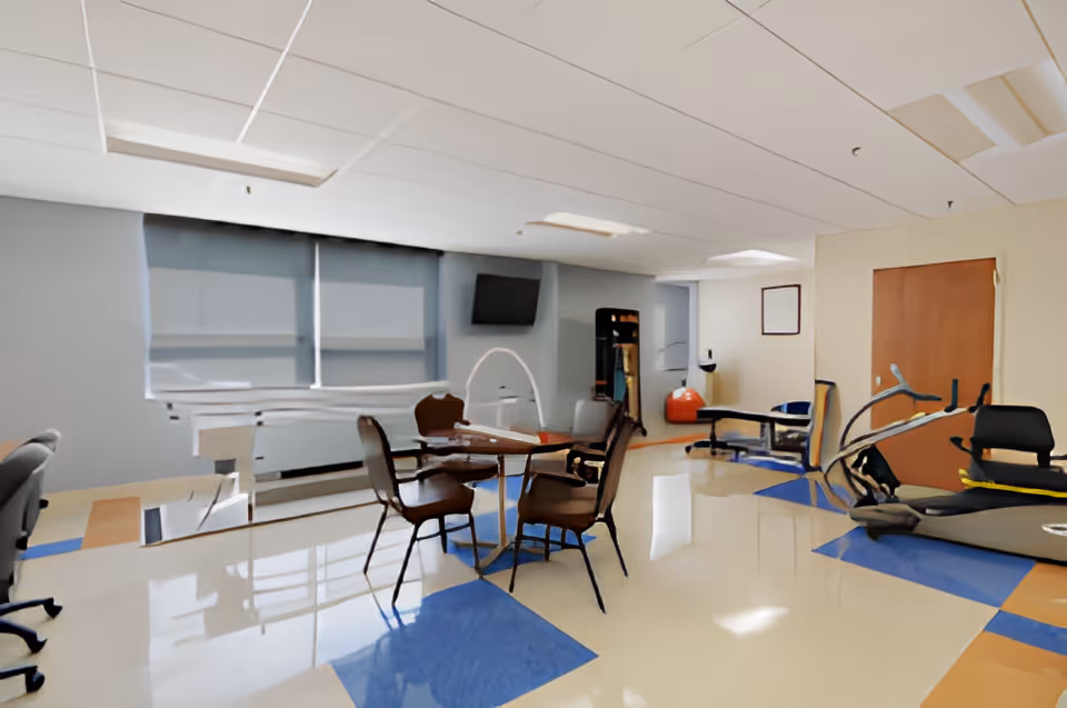 A spacious room with a table and four chairs in the center, exercise equipment including a treadmill and a stationary bike on the right, a therapy table in the back, a television mounted on the wall, and large windows with blinds partially closed.