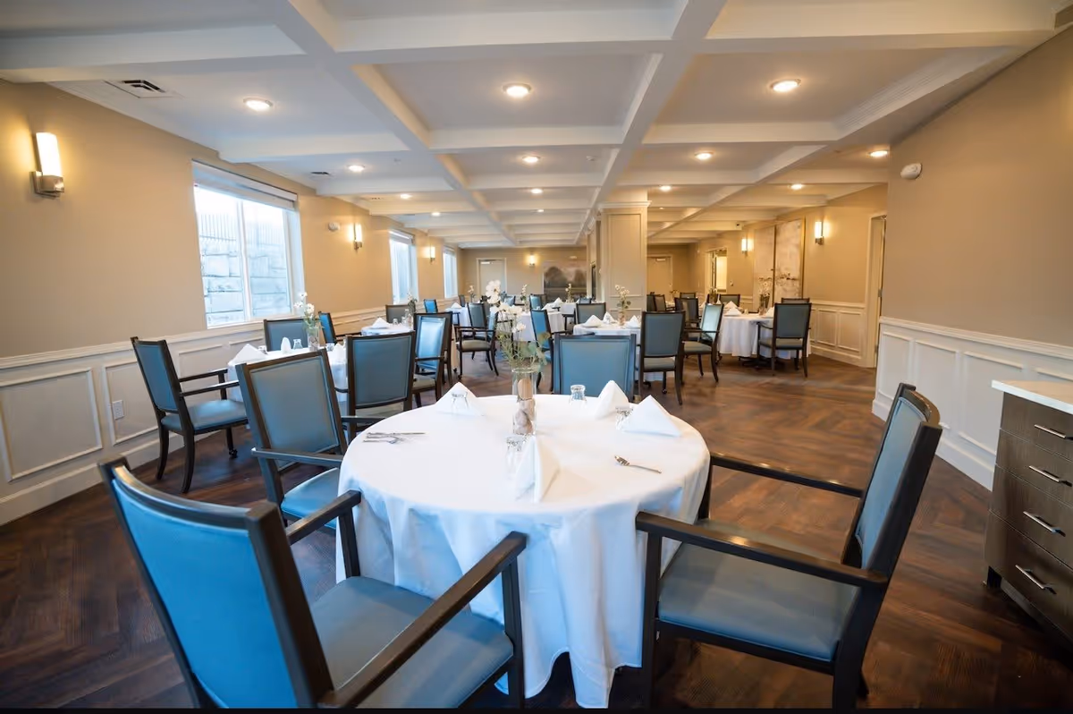 A spacious dining room in Saratoga View Senior Living with multiple round tables covered in white tablecloths, each set with folded napkins, glasses, and silverware. The room features blue cushioned chairs, wooden flooring, beige walls with white wainscoting, and ceiling lights in a coffered ceiling design. Windows along one wall allow natural light to enter.