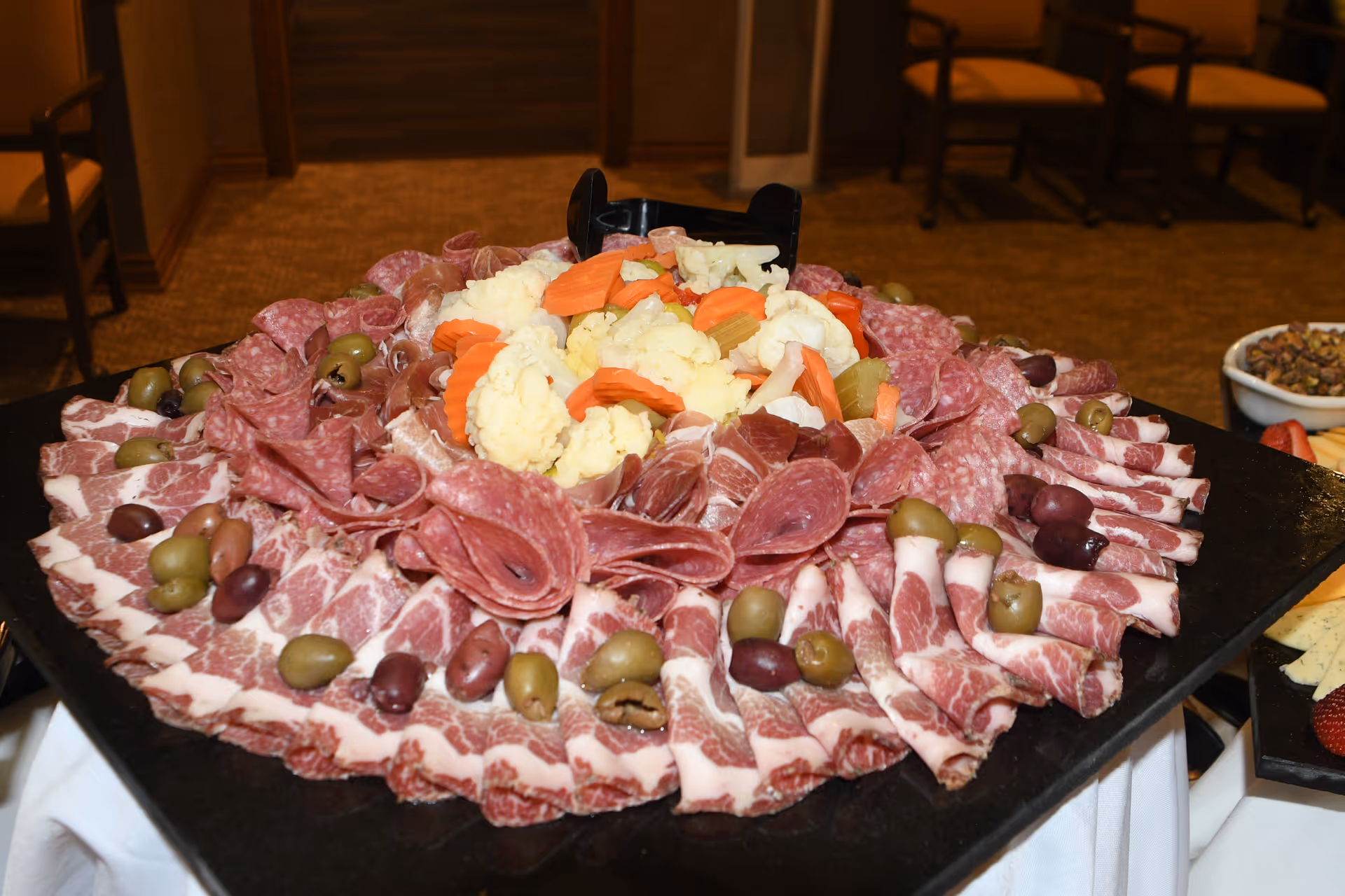 A large platter of assorted deli meats arranged in a circular pattern with green and black olives, and a center pile of pickled vegetables including cauliflower and carrots, set on a table in a room with carpeted floor and chairs in the background.